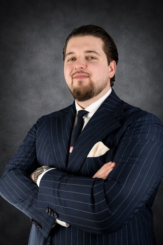 Professional portrait of a man with beard wearing a navy pinstripe suit and tie, arms crossed, against a gray background