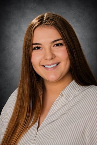 Portrait of a young woman with long brown hair, smiling at the camera, wearing a light striped shirt against a gray background